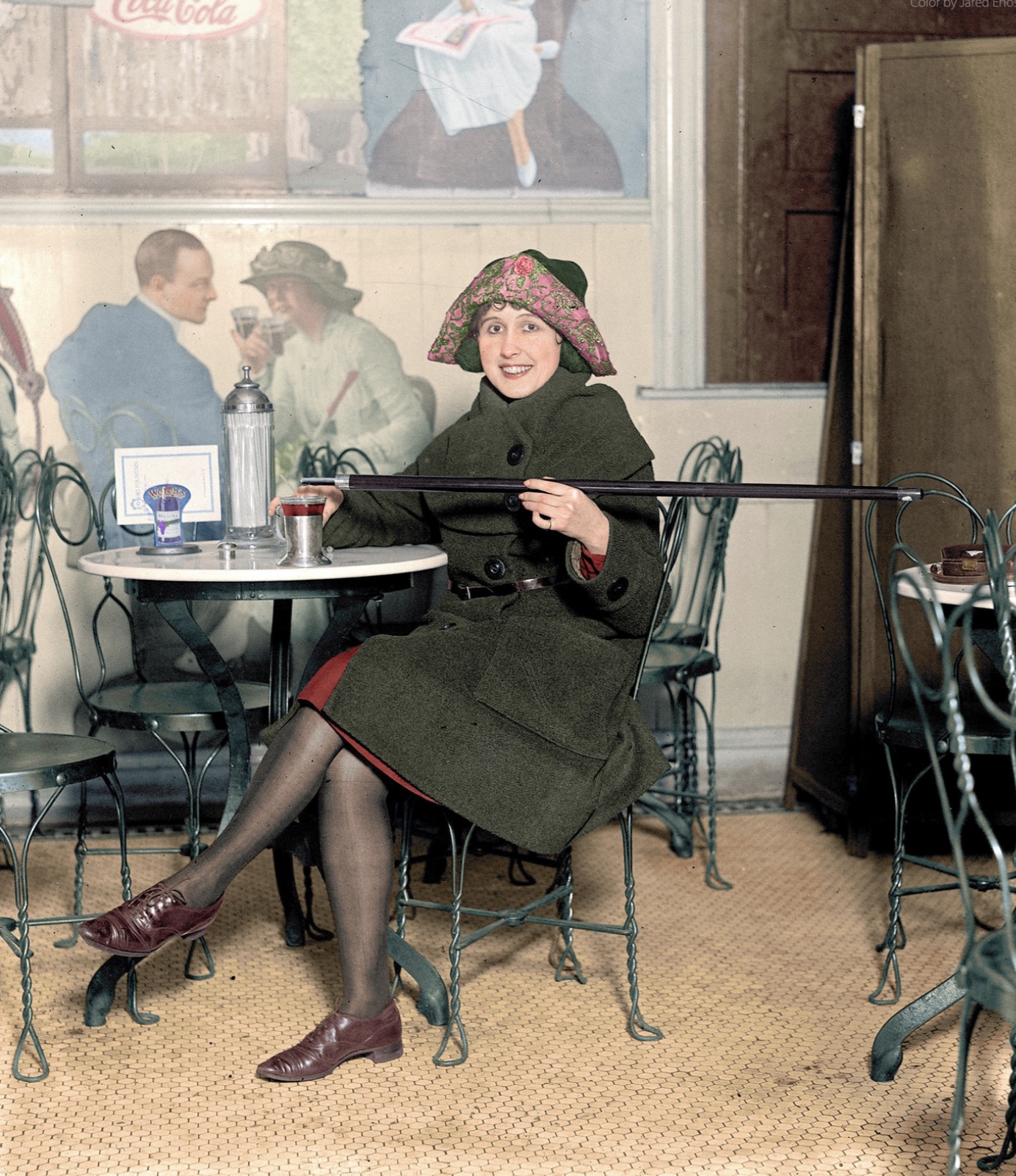 Woman seated at a soda fountain table is pouring alcohol into a cup from a cane, during Prohibition; with a large Coca-Cola advertisement on the wall, 1922 
